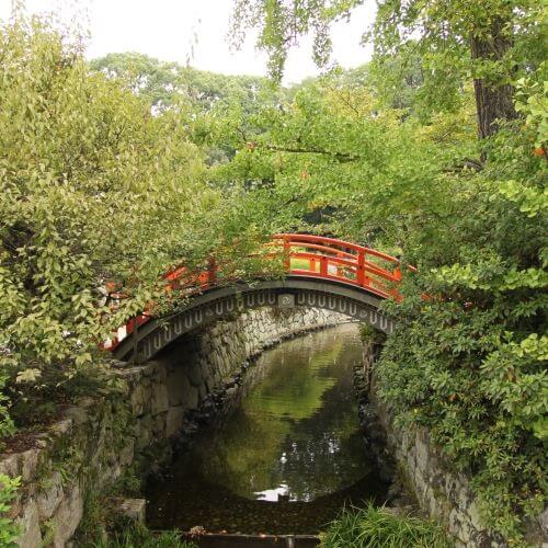 A small wooden Japanese red bridge arches over a canal surrounded by green vegetation.